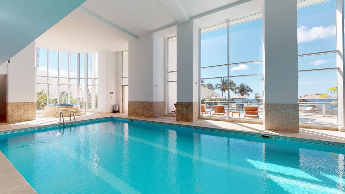 An indoor pool is visible, featuring clear blue water under bright natural light from large windows. The area is adorned with minimalist decor and has a ladder for entry. Poolside seating can be seen through the glass, offering views of the outdoors.