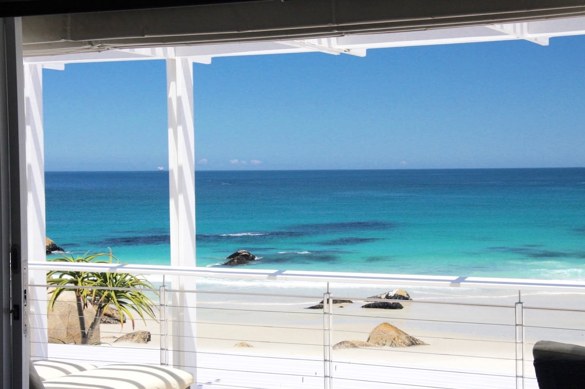 A panoramic view captures the serene ocean with shades of turquoise and deep blue, contrasting with the sandy beach below. The image showcases a clear sky, allowing natural light to filter through the open space, while rocky outcrops can be seen along the shoreline.