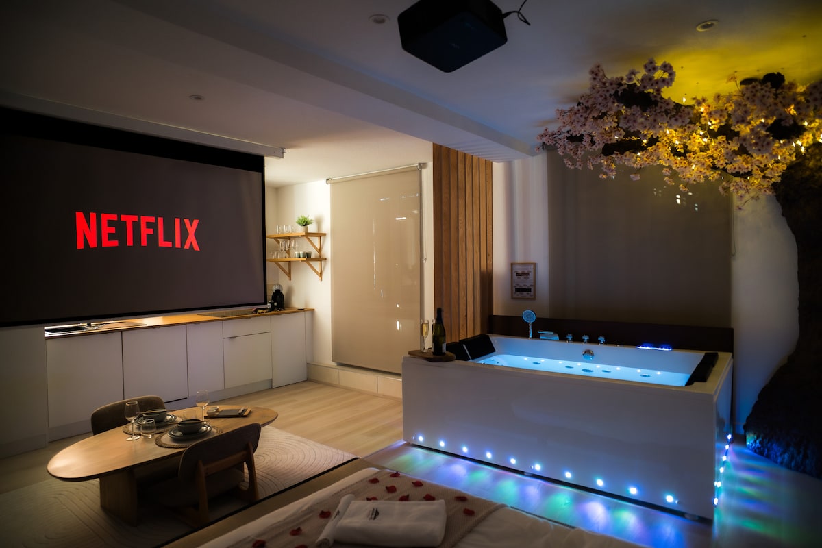 A spacious living area features a large projection screen displaying 'NETFLIX'. A hydrotherapy bath with ambient lighting is situated nearby, along with a dining table set on the floor. Soft floral decorations enhance the serene environment.