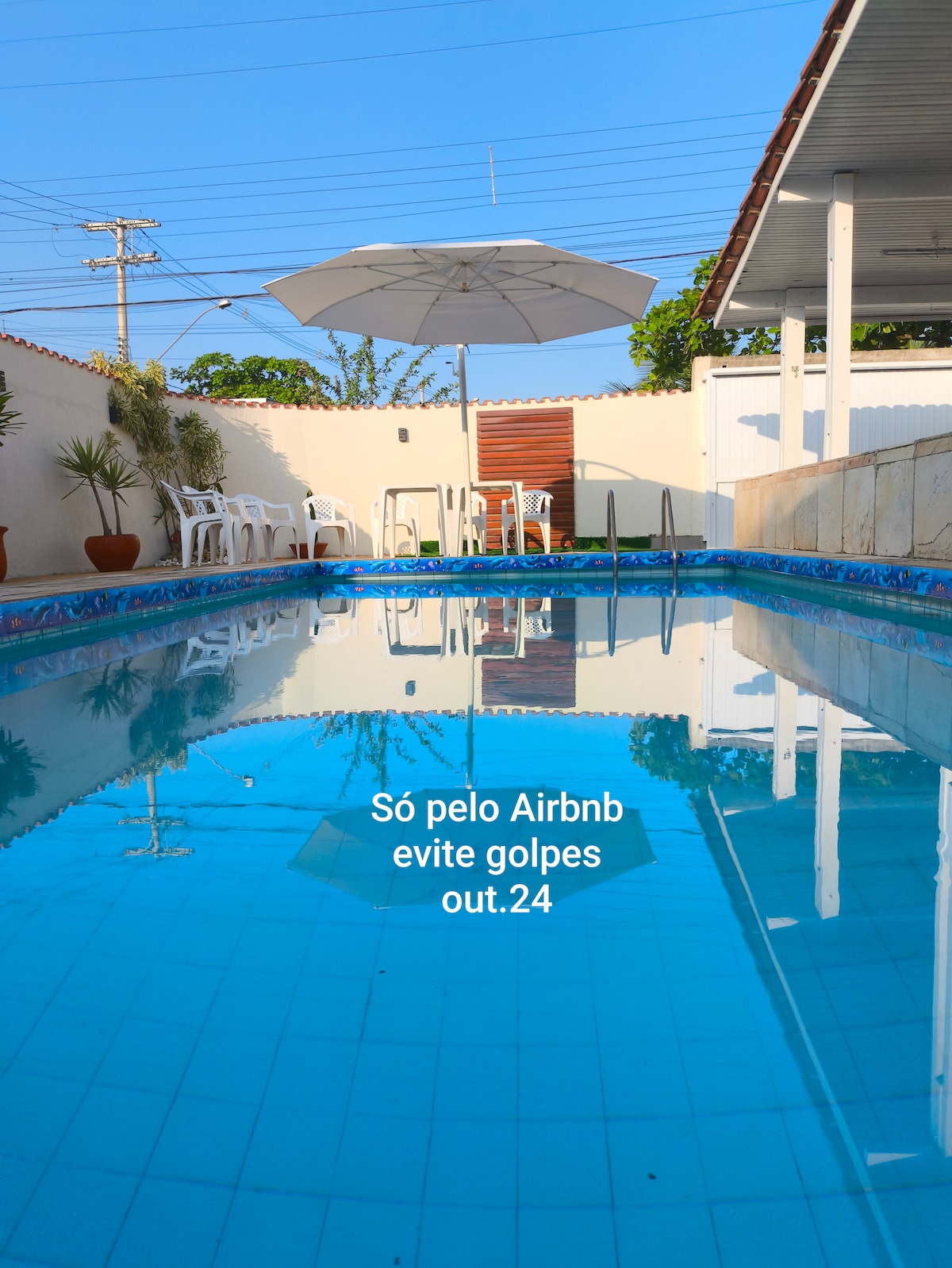 A serene swimming pool is featured under a clear blue sky, surrounded by white lounge chairs and a large umbrella for shade. The pool's water reflects the bright colors of the surroundings, creating a tranquil atmosphere for relaxation.