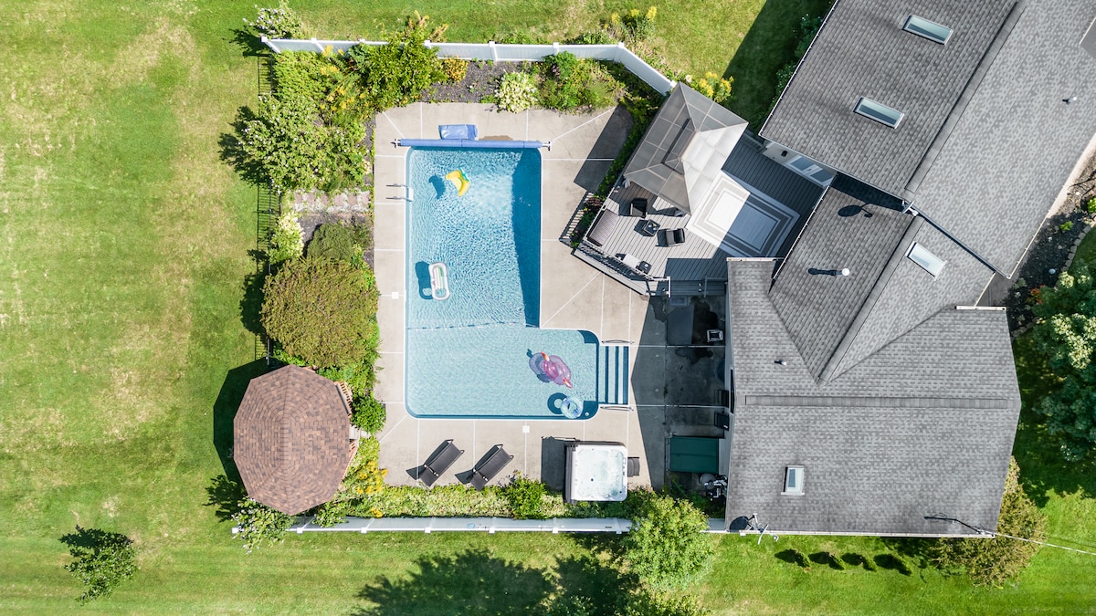 An aerial view showcases a private pool surrounded by neatly landscaped greenery. A gazebo and patio area are visible beside the pool, equipped with lounge chairs and an umbrella for shade. The residential house features a gray roof and is positioned adjacent to the pool area.
