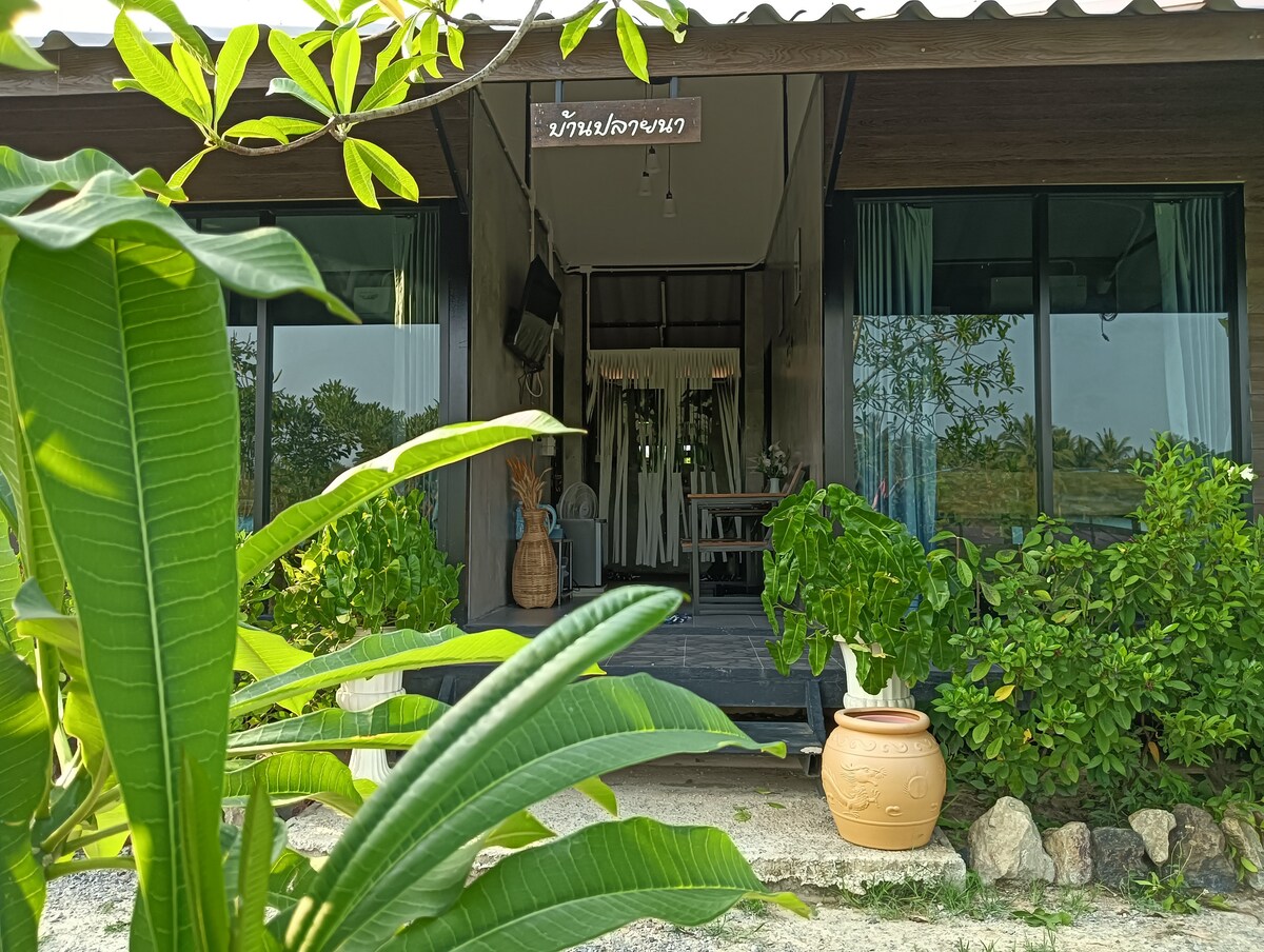 A welcoming entrance is framed by lush greenery and potted plants. Large glass windows provide views into the interior space, while a rustic sign hangs above the door. The wooden flooring leads to a cozy porch area, enhancing the connection with the surrounding landscape.