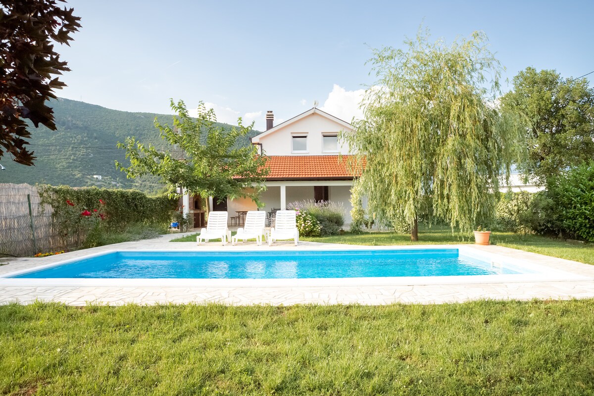 A serene garden is featured, showcasing a large outdoor swimming pool surrounded by a well-manicured lawn. Two lounge chairs are positioned beside the pool, inviting relaxation. In the background, a two-story house is visible, complemented by trees and vibrant greenery.