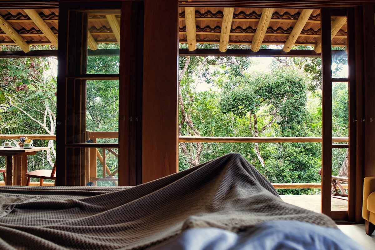 A cozy sleeping area is visible, featuring a textured blanket draped over a spacious bed. Large glass doors open to a private balcony, offering a view of the lush greenery. Natural wood elements frame the scene, enhancing the connection to the surrounding forest.