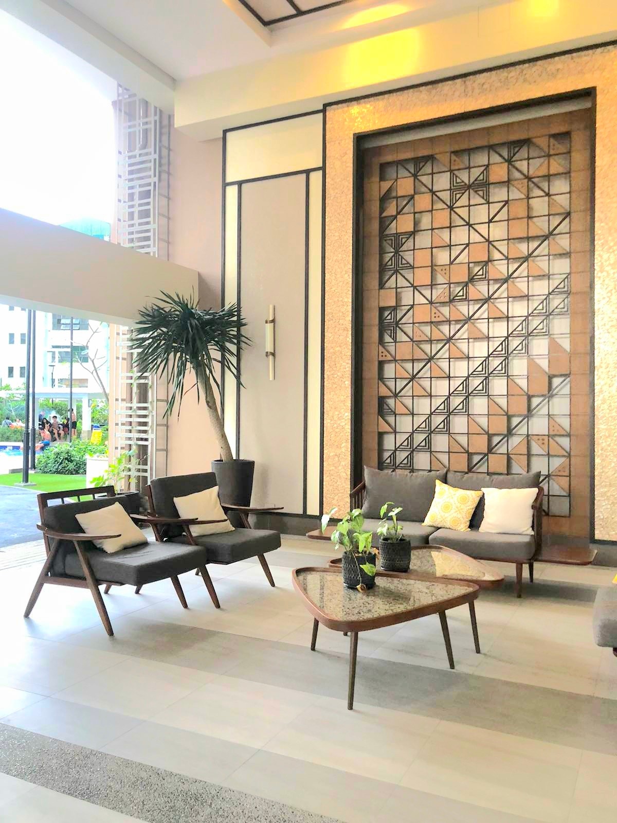 A modern lobby area is presented with a textured wall featuring geometric patterns. Comfortable seating is arranged around a low, decorative table, surrounded by potted plants. Natural light filters in through large glass doors, enhancing the inviting atmosphere of the space.