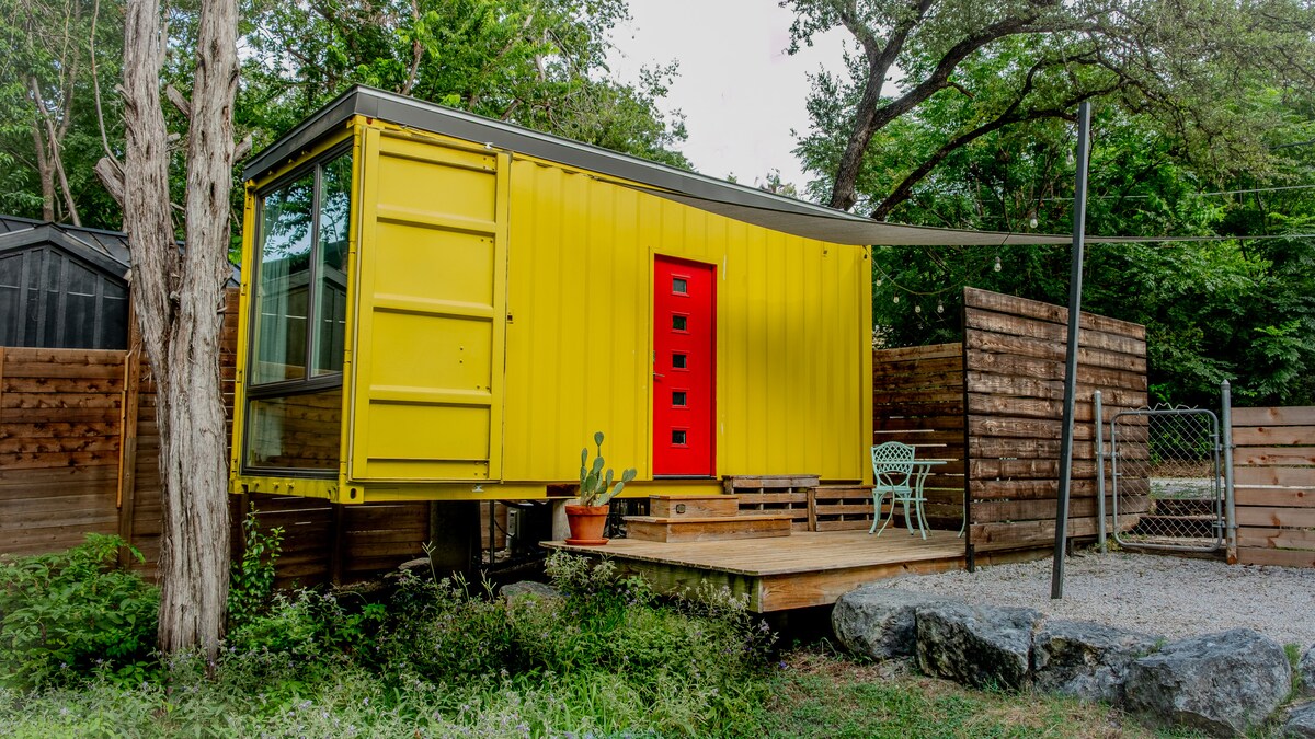 A brightly colored yellow shipping container forms the exterior of the accommodation, complemented by a striking red door and matching accents. A small wooden deck leads to the entrance, bordered by landscaping that includes stones and greenery. A bistro table and chairs are arranged on the patio.