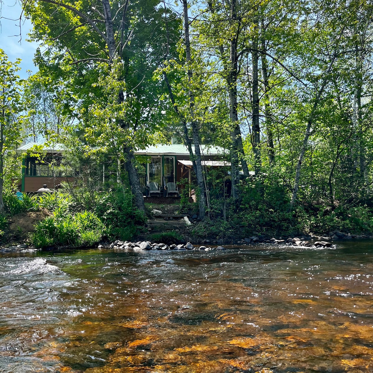 A serene view of a chalet located beside the Mastigouche River, surrounded by lush greenery. The structure features large windows, inviting natural light, and is complemented by the gentle flow of the river nearby.