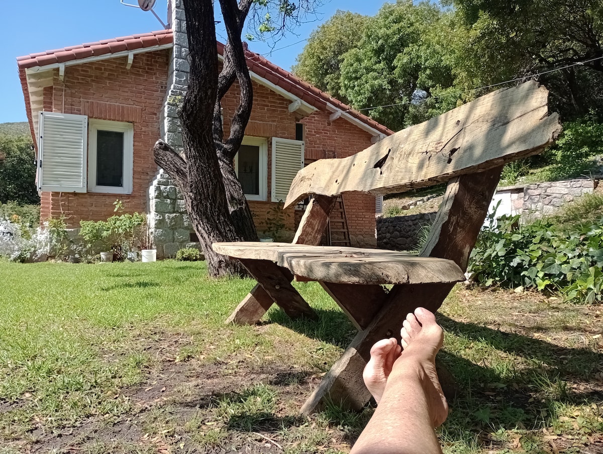 A rustic wooden bench is positioned in a grassy area, offering a view of a cozy brick cottage with white shutters. Lush greenery surrounds the setting, including trees and plants, creating a peaceful atmosphere. The ground is shown with bare feet relaxing in the foreground.
