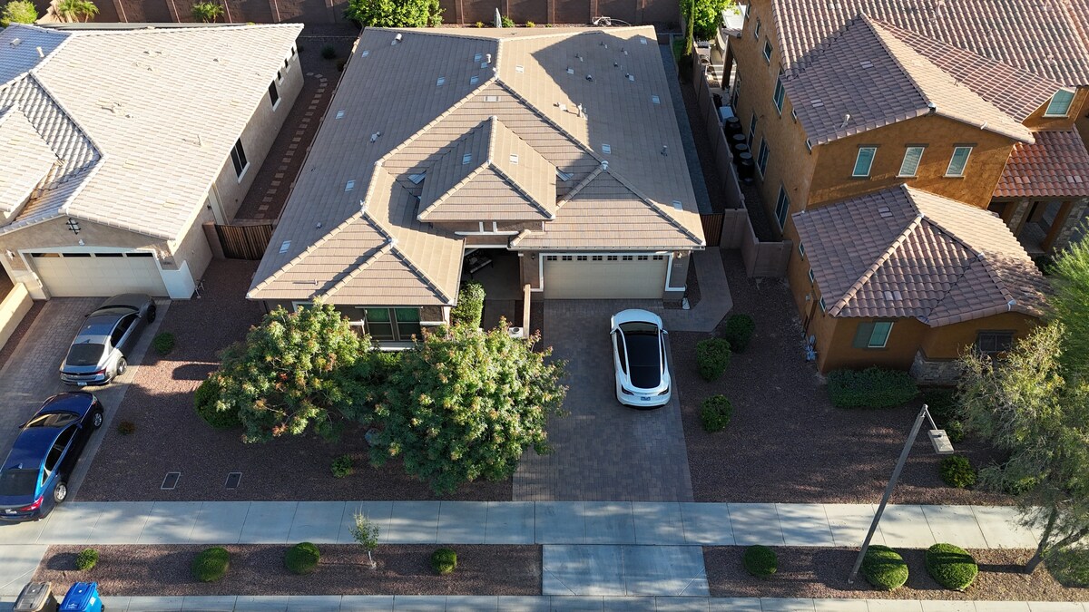 An aerial view of a residential area showing multiple homes. A driveway with two parked cars is visible, surrounded by neatly trimmed shrubs and gravel landscaping. The garage doors are closed, and a paved pathway runs alongside the houses.