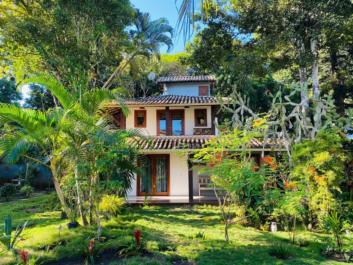 The exterior of a two-story house is surrounded by lush greenery and colorful plants. Large windows reflect natural light, and a tiled roof adds character to the structure. The peaceful garden space features well-maintained grass and a variety of tropical foliage.