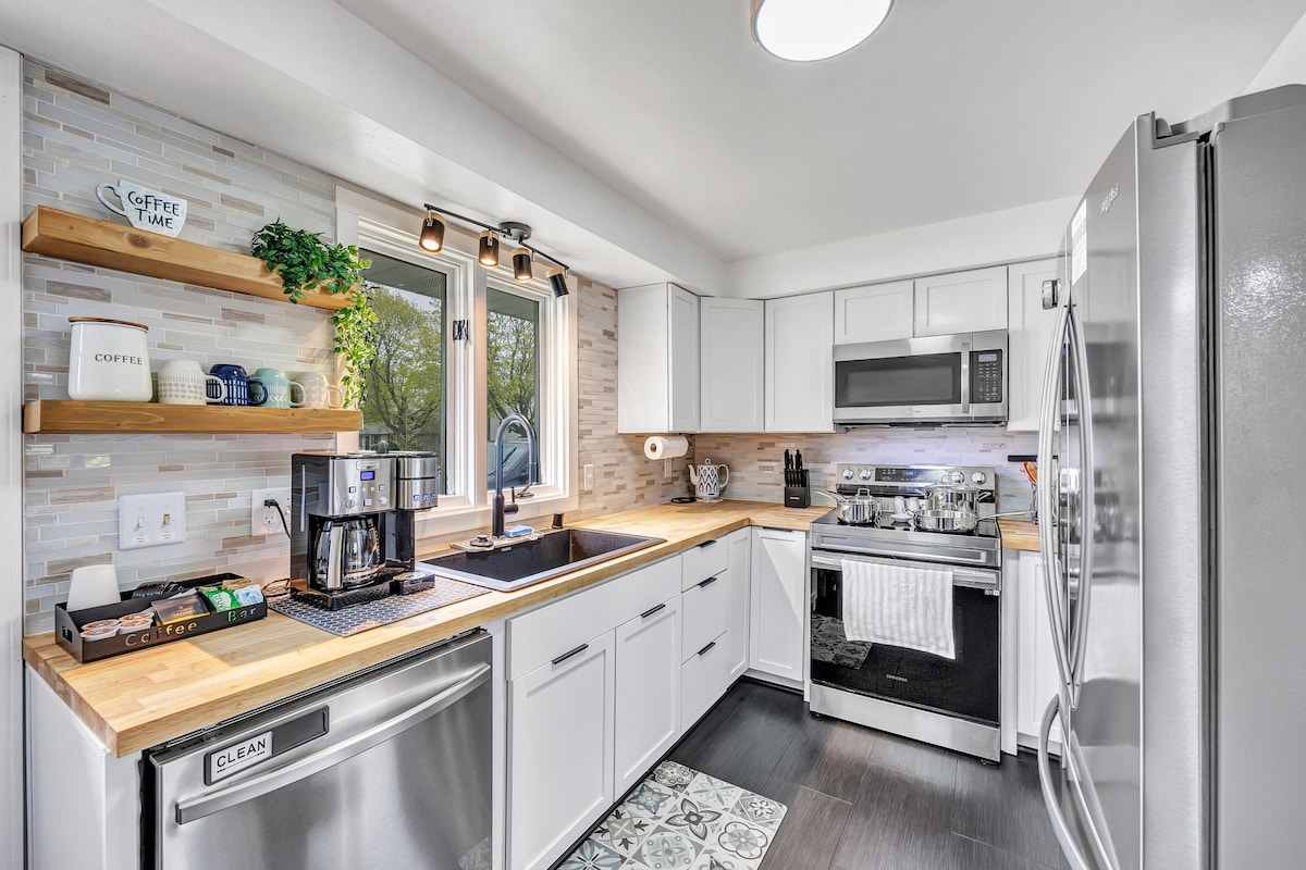 A modern kitchen features sleek white cabinetry and light wood countertops. Stainless steel appliances include a refrigerator and microwave. A coffee station with various brewing options is situated near a window that provides natural light. Decorative shelves hold coffee mugs and decorative items.
