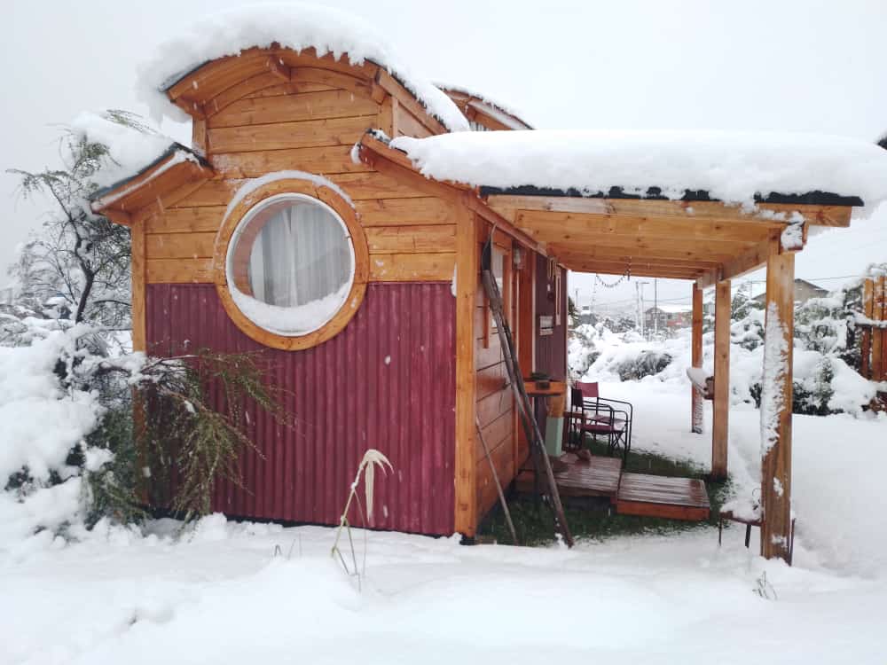 A charming tiny house is surrounded by a blanket of snow, showcasing wooden exterior features. A round window is prominently displayed, while a porch area provides access to the outdoors. Snow-covered plants and a few outdoor chairs accentuate the tranquil winter setting.