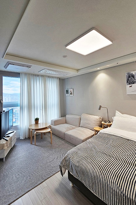 A well-organized room features a spacious bed with a striped blanket and a cozy sitting area with a light-colored sofa. A small round table complements the space, while large windows allow natural light to fill the room, offering glimpses of the cityscape outside.