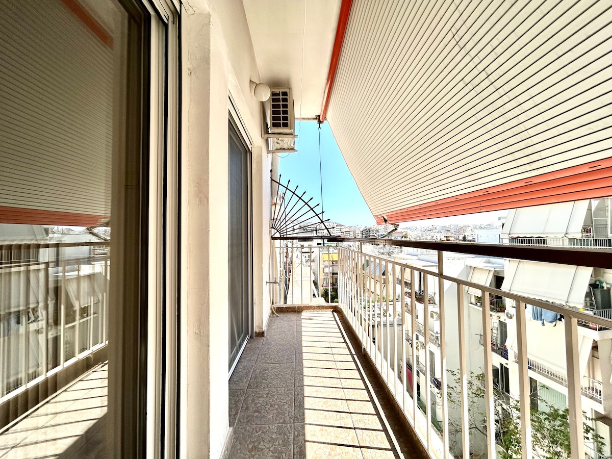 A rear balcony is visible, featuring a railing and a tiled floor. Sunlight casts shadows from the surrounding structures. A view of the courtyard and distant cityscape can be seen, with clear skies providing a bright atmosphere.