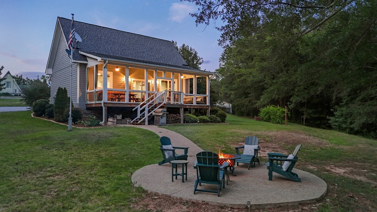 A charming house is set on a grassy lot, featuring a welcoming front porch and pathways leading to an outdoor fire pit area. Surrounding the fire pit are several Adirondack chairs, inviting relaxation. The space is bordered by trees, enhancing the serene atmosphere.