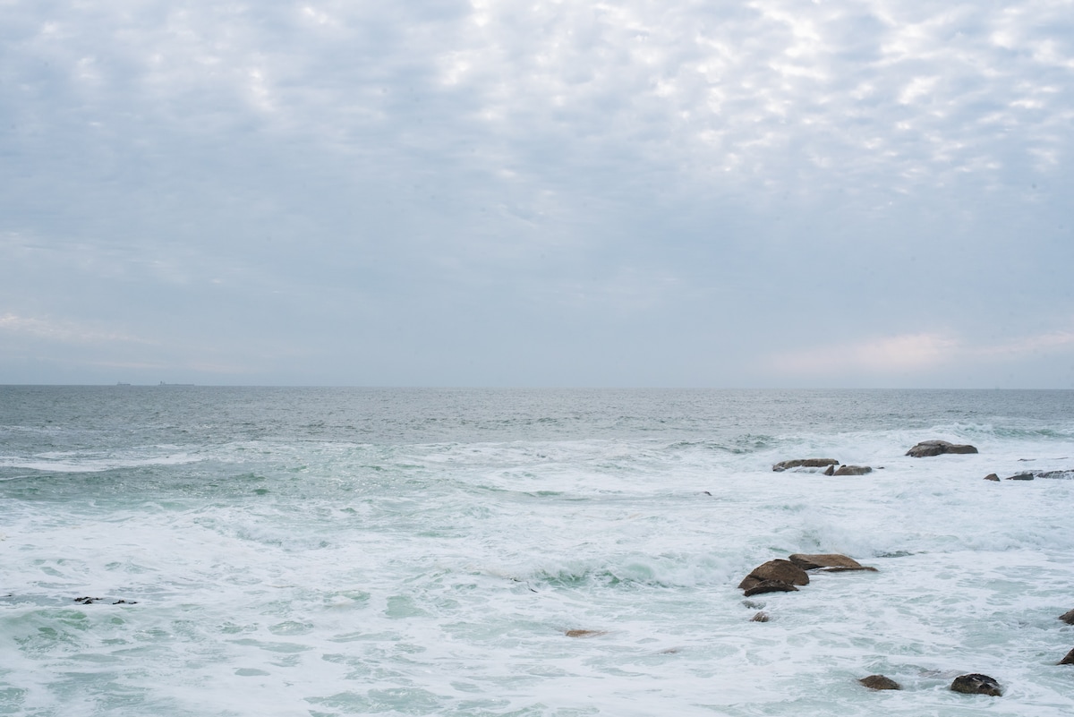 A serene view of the ocean stretches across the horizon, with gentle waves creating a rhythmic motion against the rocky shoreline. The sky is filled with soft clouds, blending shades of grey and blue, reflecting the calm atmosphere of the coastal scene.