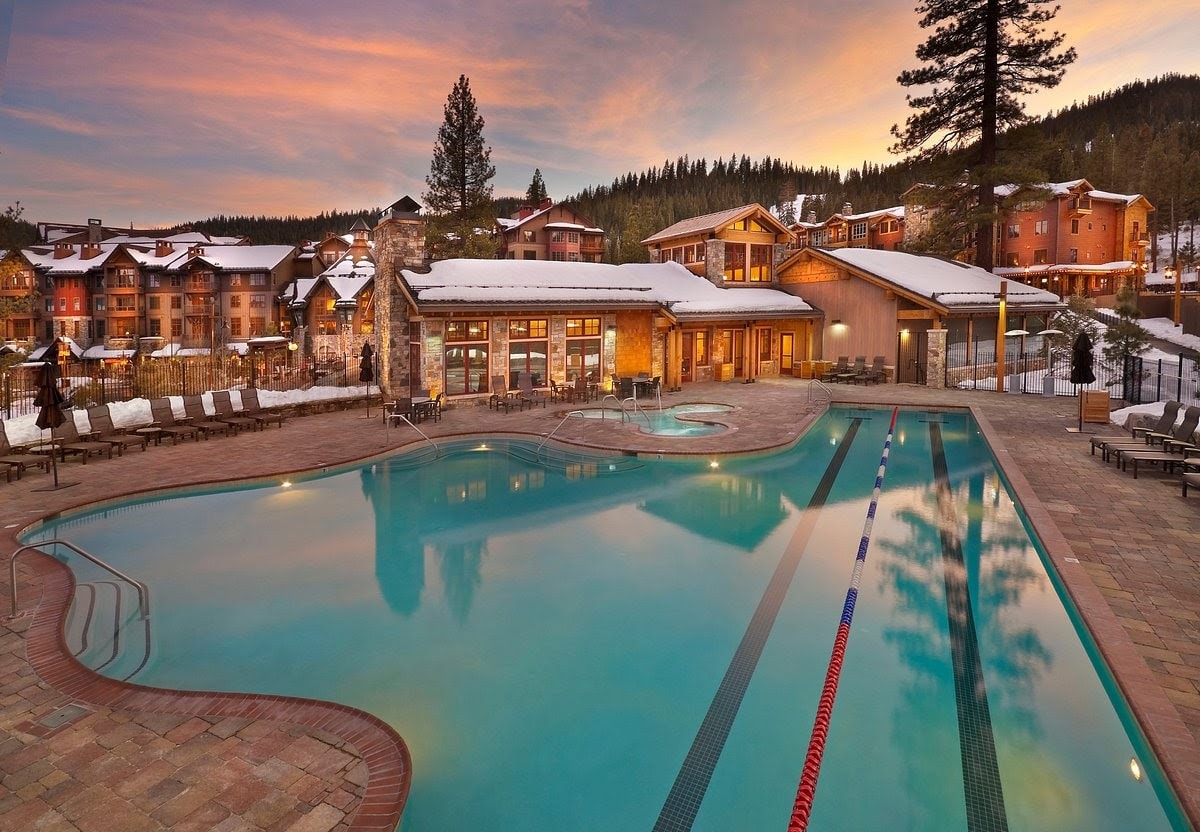 An outdoor heated pool is surrounded by stone decking and lounge chairs, with a hot tub nearby. The setting is enhanced by snow-capped trees and mountains in the background, while the sunset casts soft colors across the sky, reflecting on the water's surface.