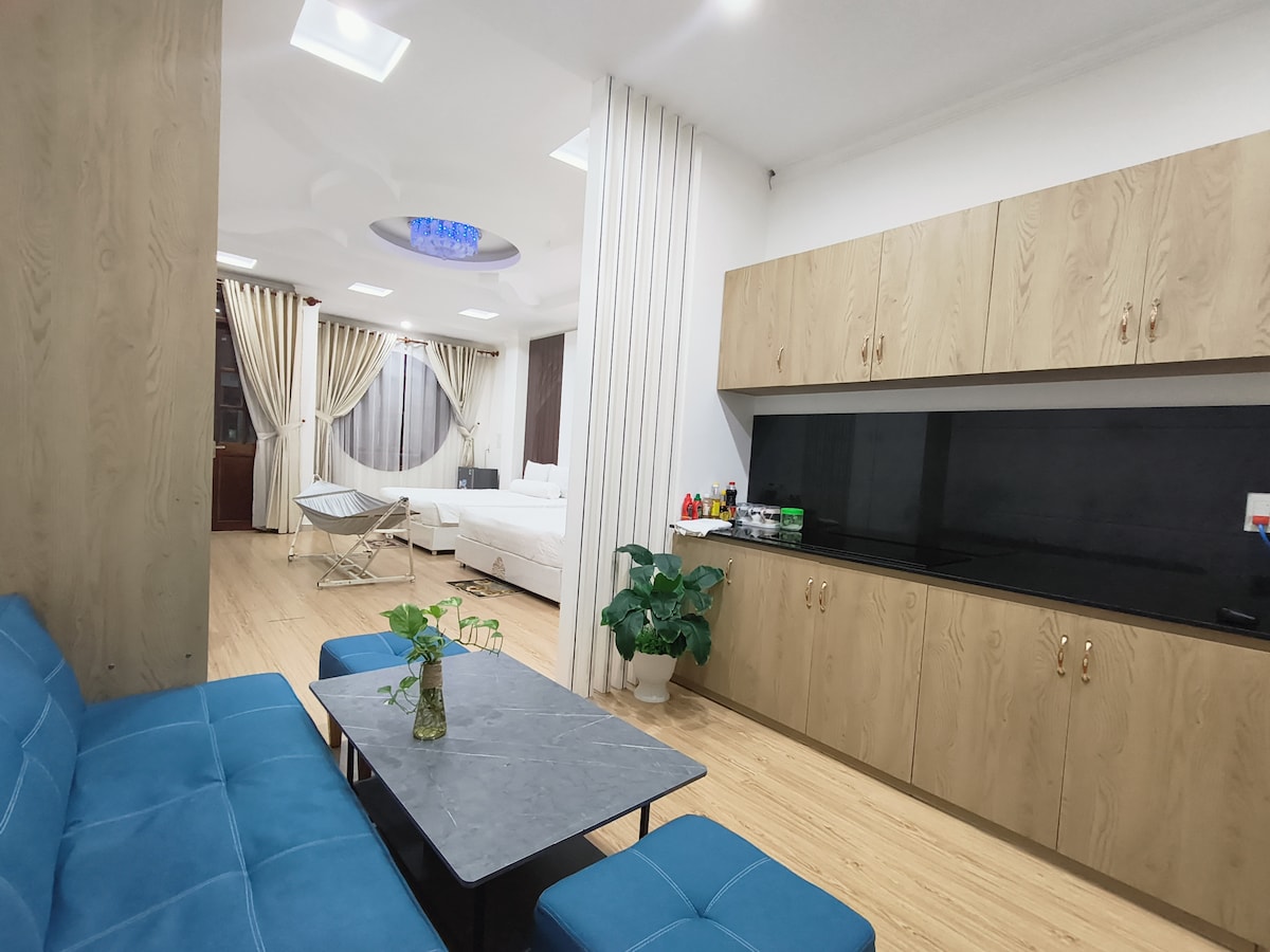A spacious living area is presented with a blue sofa and a marble-top table. A kitchenette with wood cabinetry and a dark countertop is visible, along with a green plant. Natural light enters through large windows dressed in light curtains, leading to a sleeping area with a bed.