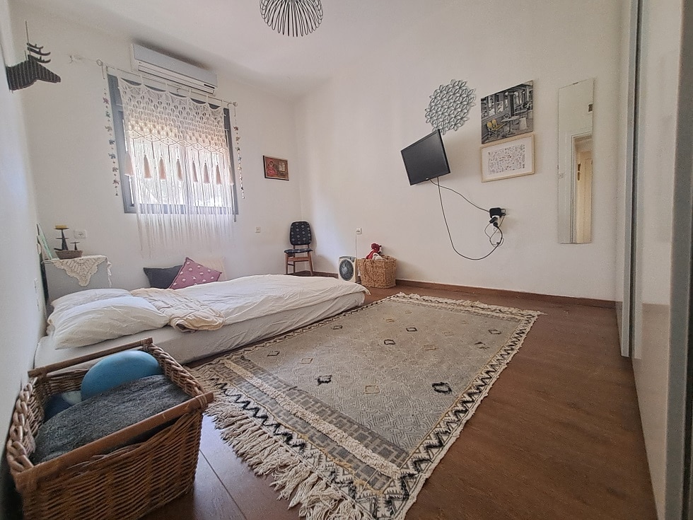 The guest room features two spacious mattresses placed on a wooden floor, surrounded by a patterned area rug. A mounted TV and wall decor, including framed art and a mirror, add character. Natural light floods through a window, and air conditioning is visible.