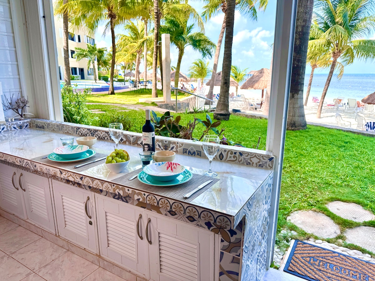 A welcoming dining area features a marble countertop adorned with plates and wine glasses, set for a meal. Through large glass doors, views of a lush garden with palm trees and a sandy beach are visible, creating a serene backdrop.