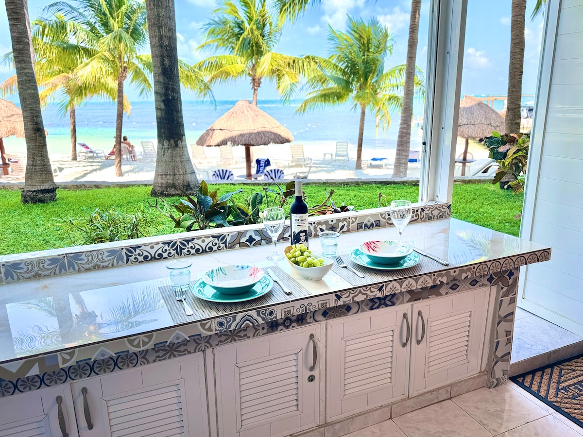 A kitchen counter is positioned by large windows, offering a view of palm trees and the beach. Plates are set for two, complemented by a bottle of wine and a bowl of grapes. The serene beach scenery enhances the inviting dining space.