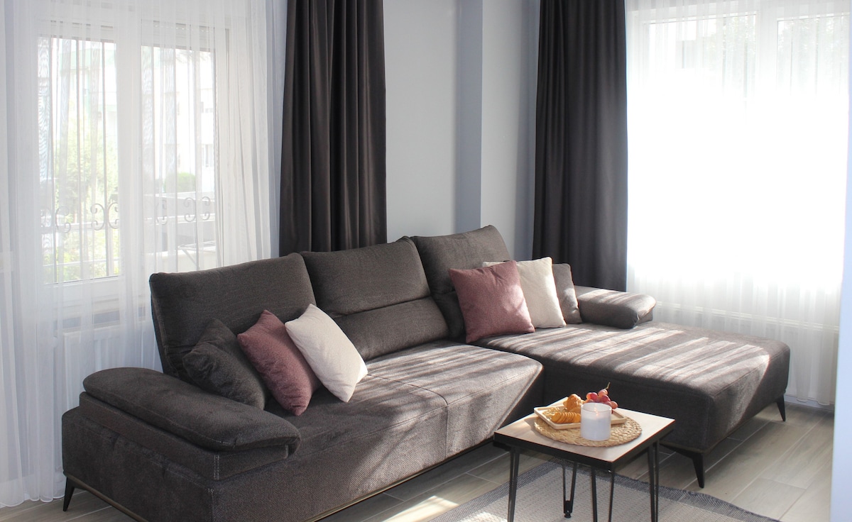 A spacious living area features a large, dark grey sectional sofa adorned with several cushions in soft tones. Natural light filters through sheer curtains, illuminating the room. A simple coffee table sits in front of the sofa, accompanied by light snacks displayed in an inviting manner.
