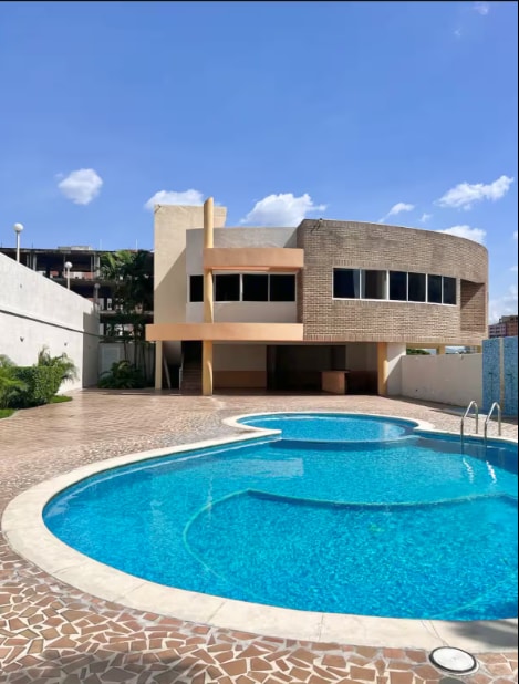 A swimming pool with a rounded shape is surrounded by a spacious deck made of patterned tiles. A two-story building with large windows is positioned beside the pool, featuring an exterior of brick and stucco. Clear blue skies and fluffy clouds complement the scene.