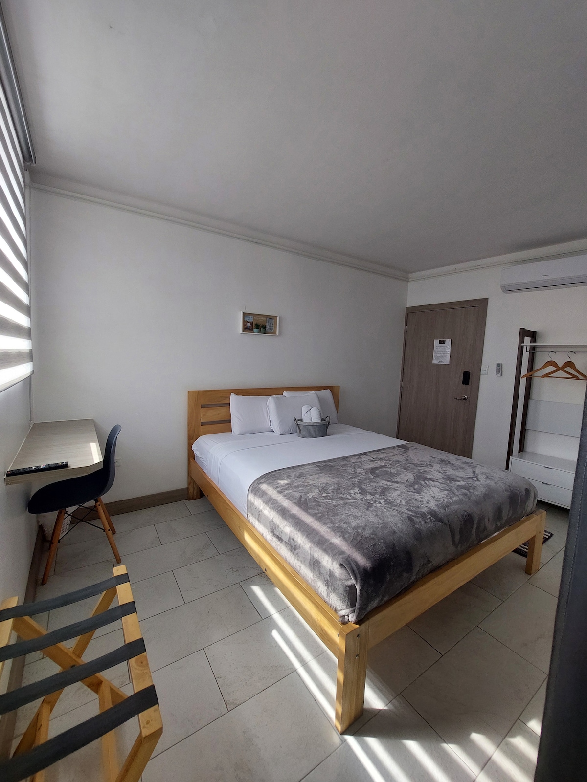 A modern bedroom features a spacious bed adorned with neutral bedding. A sleek desk and chair are positioned nearby, alongside a minimalistic clothing rack. Natural light filters through partially closed blinds, illuminating the light-colored walls and tiled floor.