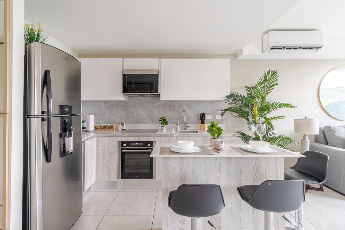 The kitchen features modern appliances, including a black refrigerator and built-in oven. A small island with sleek stools offers additional seating. A plant adds a touch of greenery, while the light countertops contrast with the dark cabinetry, creating an inviting cooking space.