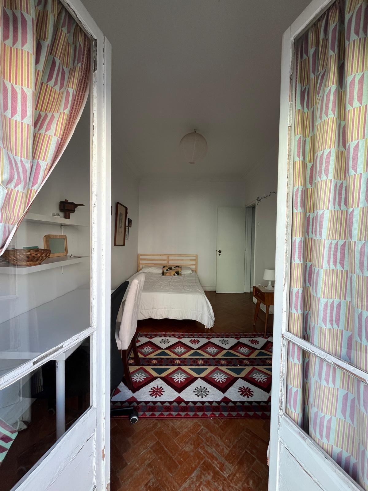 A glimpse into the private room reveals a light-colored bed positioned against a white wall, framed by patterned curtains. A decorative rug adds warmth to the wooden floor. The space is illuminated by natural light, contributing to a serene atmosphere.