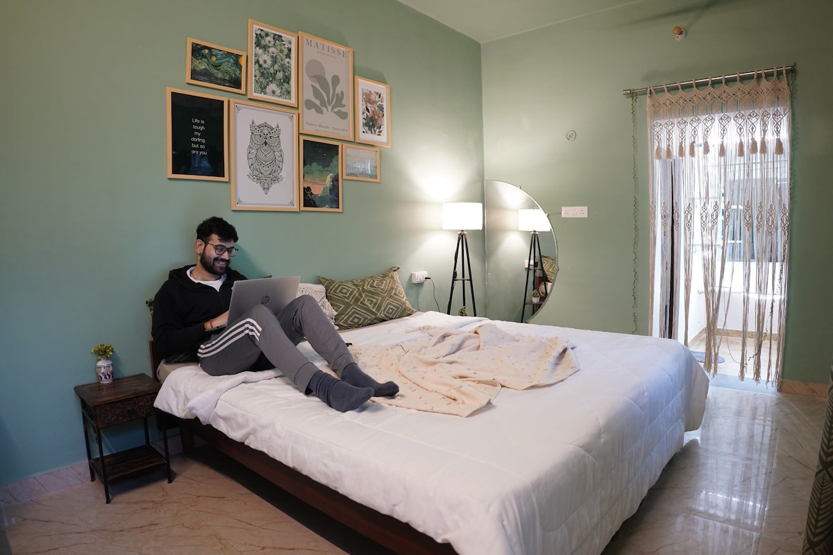A cozy bedroom features dry sage green walls adorned with framed artwork. A spacious bed with a light blanket is positioned next to a nightstand, while a person is seated on the bed using a laptop. Natural light enters through a window, complemented by a floor lamp.