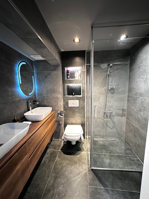 The modern bathroom features a sleek design with a glass shower enclosure and a wall-mounted toilet. Dual sinks are set atop a wooden vanity, and ambient lighting highlights the contemporary finishes. A circular mirror reflects the space, enhancing the overall aesthetic.