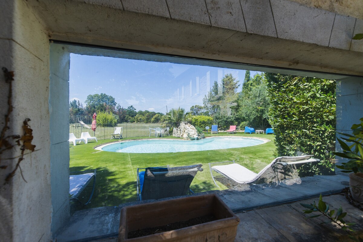 A large window frames a view of a swimming pool surrounded by manicured grass. Comfortable lounge chairs and sunbeds are positioned around the pool, offering a serene setting. Lush greenery lines the space, contributing to an inviting outdoor atmosphere.