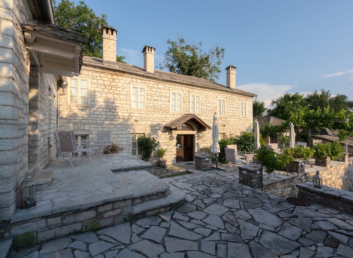 A stone building with multiple large windows and a welcoming entrance is presented, surrounded by a paved outdoor area. The space features tables and chairs, alongside sun umbrellas, inviting guests to enjoy the serene environment and views of the surrounding greenery.