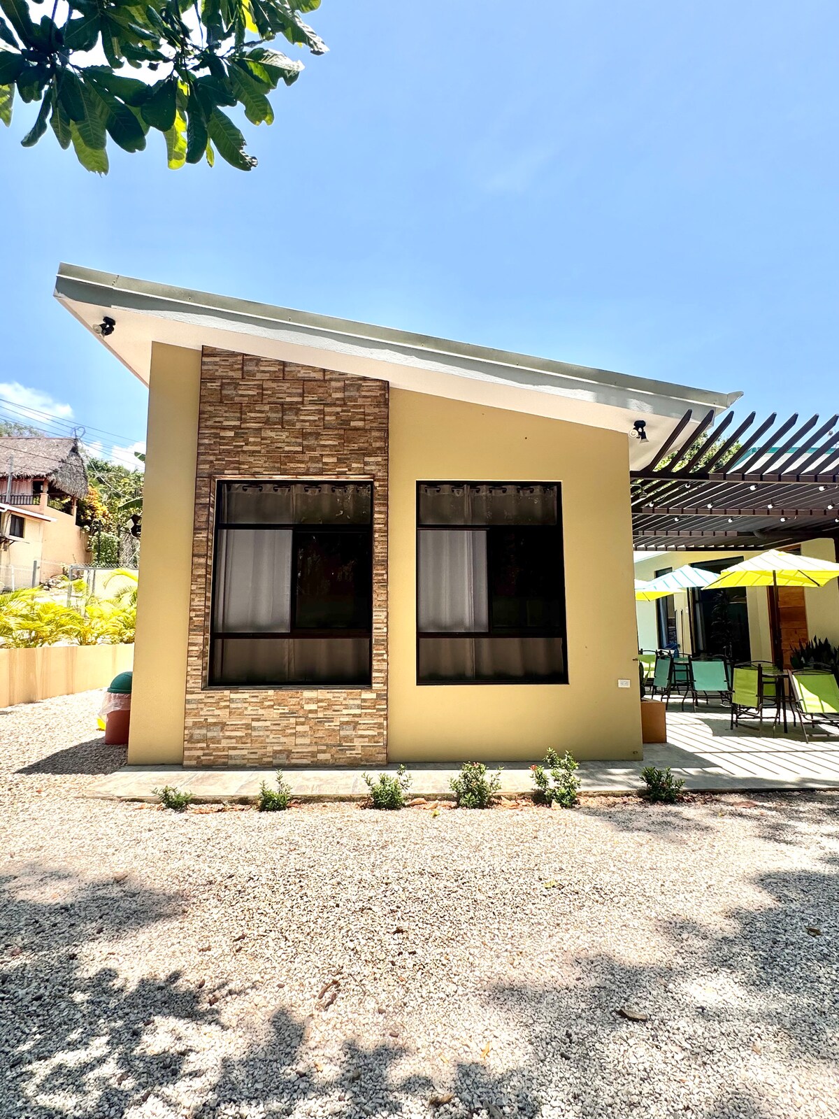 The villa's exterior is showcased, featuring a combination of textured stone and smooth wall surfaces. Large windows allow natural light to enter, framed by lush greenery. A gravel pathway leads to the entrance, where a shaded seating area with colorful umbrellas is visible.