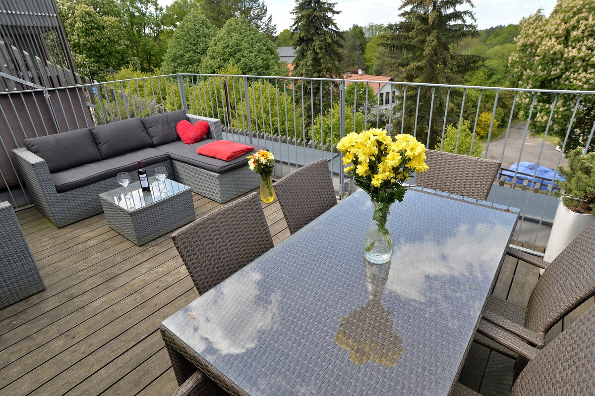 A spacious terrace is showcased with a glass-topped dining table surrounded by chairs. A comfortable seating area with a cozy couch is visible, adorned with decorative cushions. Bright yellow flowers in a vase add a touch of color, complemented by lush greenery in the background.