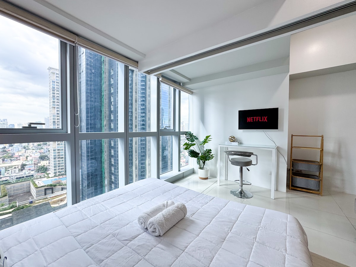 A spacious bedroom is illuminated with natural light from large floor-to-ceiling windows. A neatly arranged bed is positioned in front of the window, with a towel placed on the covers. A small chair and a TV displaying 'Netflix' are visible, adding to the cozy ambiance.