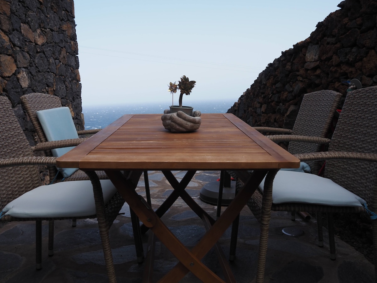A wooden outdoor dining table is present, surrounded by four wicker chairs with cushions. A decorative centerpiece featuring small plants is displayed on the table. The view in the background reveals a distant ocean and rocky landscape.