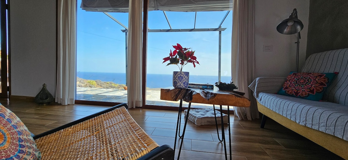 An inviting living area features a wooden coffee table with a decorative plant. Large glass doors frame a panoramic ocean view, complemented by natural light streaming into the space. A cozy seating area is adorned with colorful cushions and a comfortable sofa.