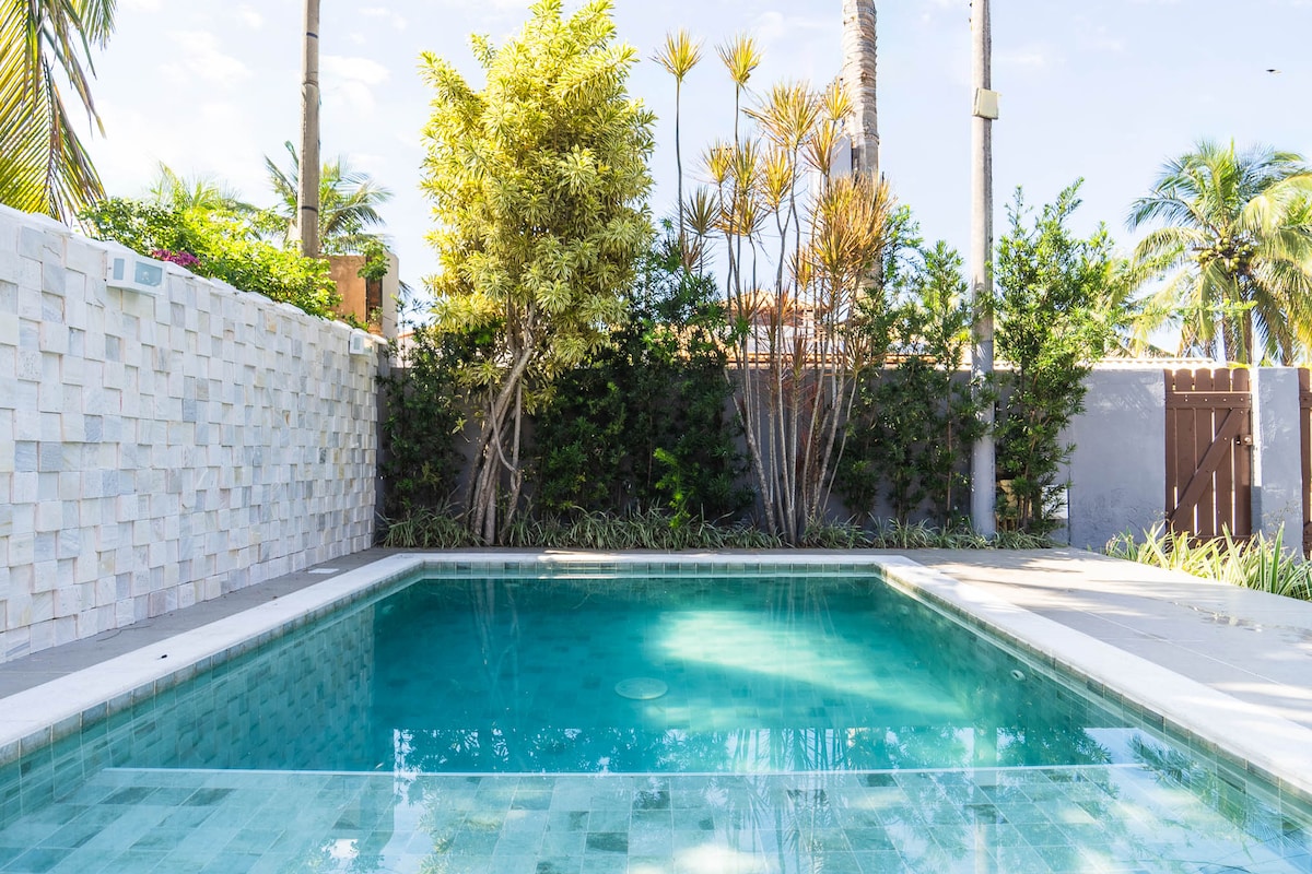 A serene pool area is framed by lush tropical plants, providing a calm ambiance. The clear water reflects the sunlight, and a stone wall adds privacy. Surrounding greenery enhances the outdoor space, ideal for relaxation and gatherings.