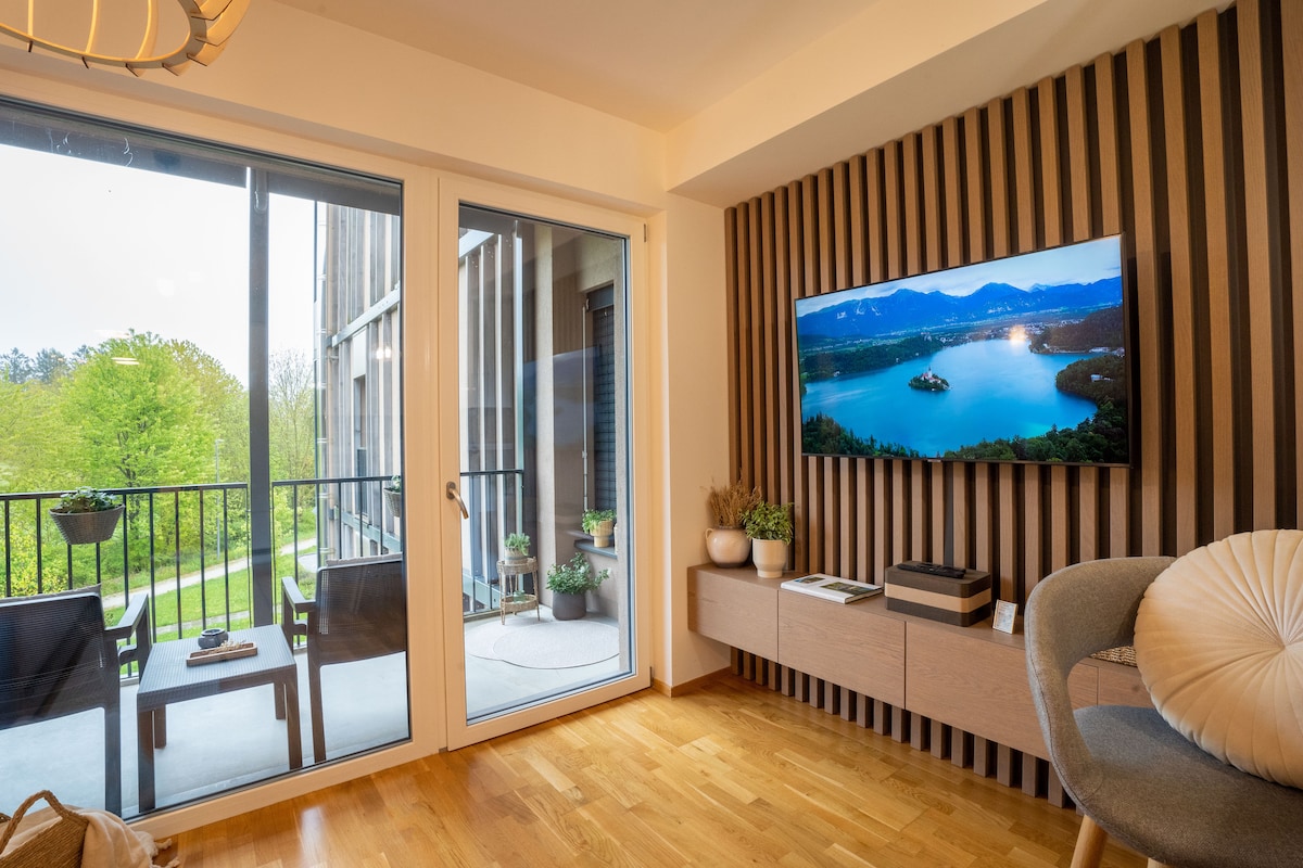 A modern living area features a mounted television showing a tranquil lakeside view. Large glass doors lead to a small balcony with a seating arrangement. Natural light fills the space, highlighting wood accents and a stylish chair placed within the inviting interior.