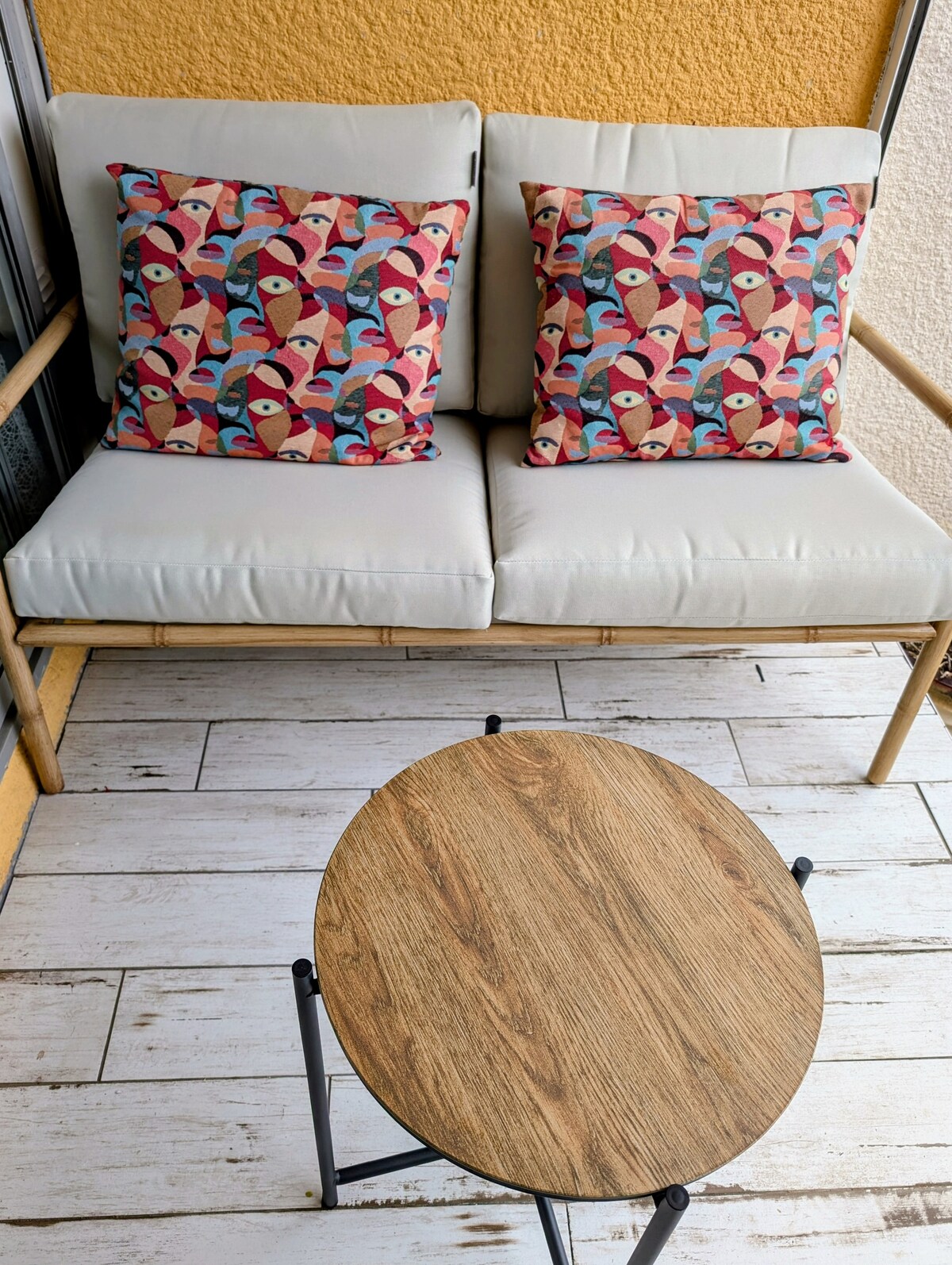 A two-seater sofa with a light-colored cushion is positioned beside a round wooden table. The sofa is adorned with colorful patterned cushions featuring abstract designs, creating a relaxing outdoor space.