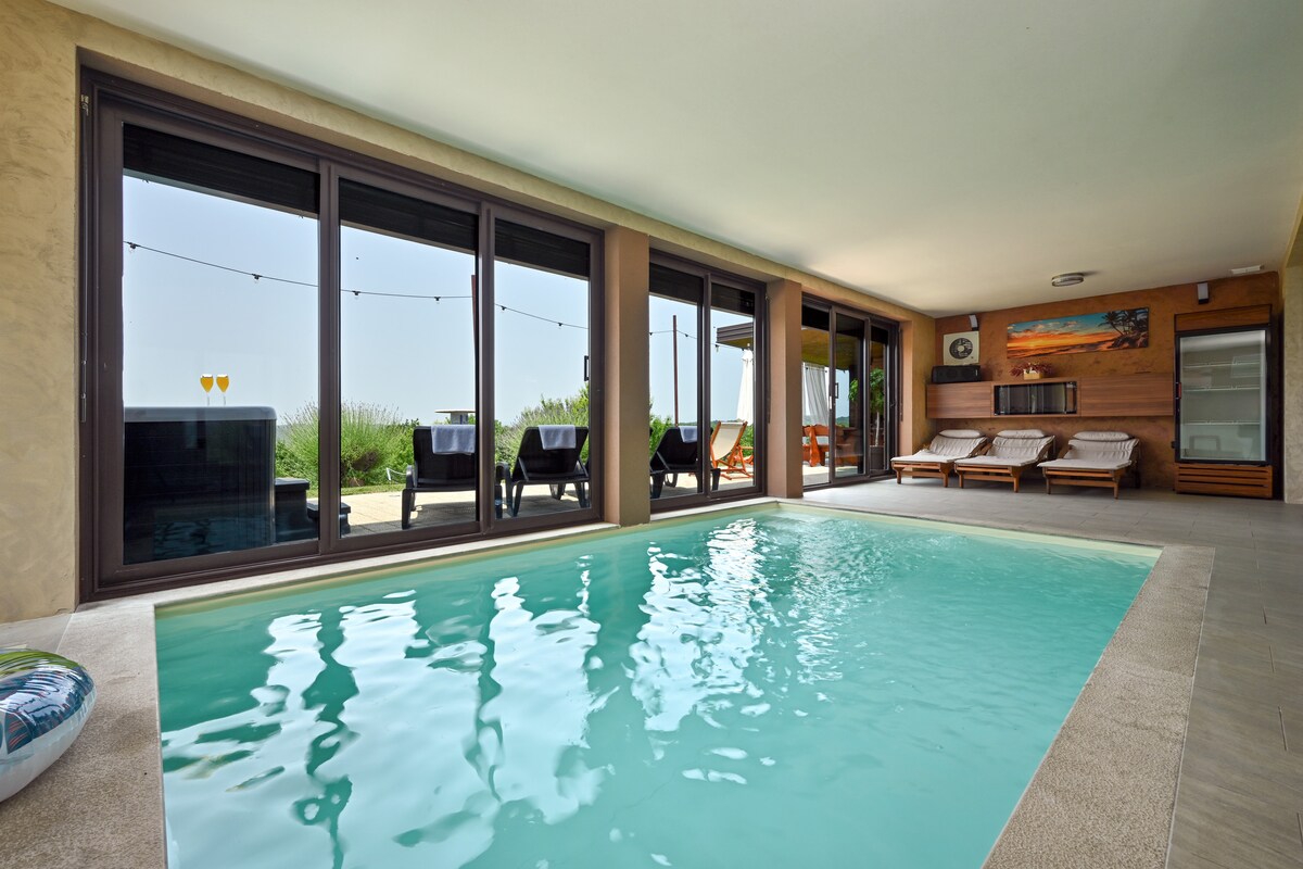 An indoor pool is showcased, framed by large glass doors that provide views of the outdoor space. Comfortable loungers are arranged beside the pool, with sunlight illuminating the serene water. The backdrop features soft greenery, enhancing the atmosphere of relaxation.