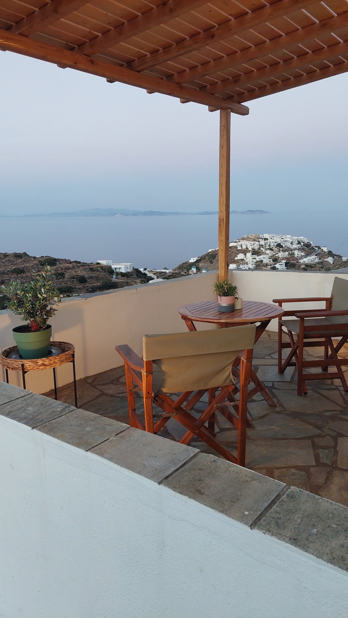 A spacious terrace is showcased with wooden furniture, including a round table and two chairs. Potted plants are placed on a small side table. An expansive view of the sea and surrounding landscape is visible, highlighting the natural beauty of the area.
