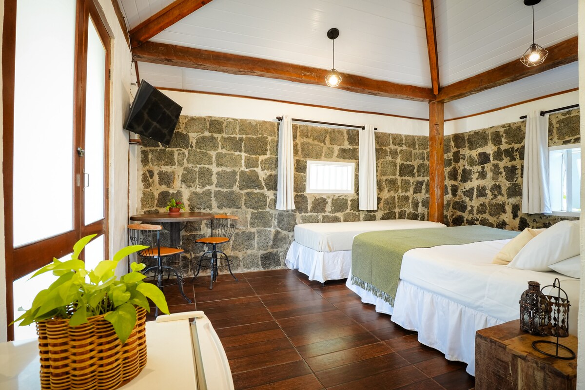 The suite features a blend of stone and wood elements, showcasing two beds with white linens and a vibrant green throw. A small dining table with two chairs is positioned near a window, while a television is mounted on the wall opposite the beds. A plant adds a touch of greenery to the space.