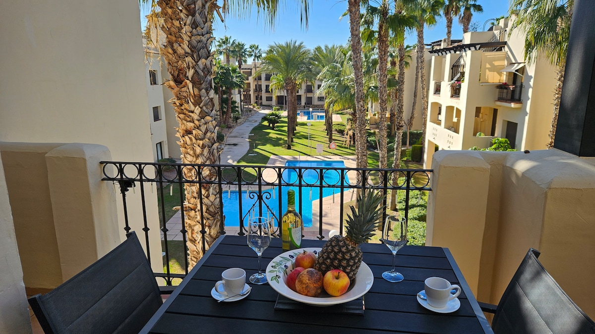 A balcony table is set with fruit and glassware, overlooking a landscaped area featuring palm trees and swimming pools. The sunny scene highlights the resort's garden design, providing a serene view of the grounds.