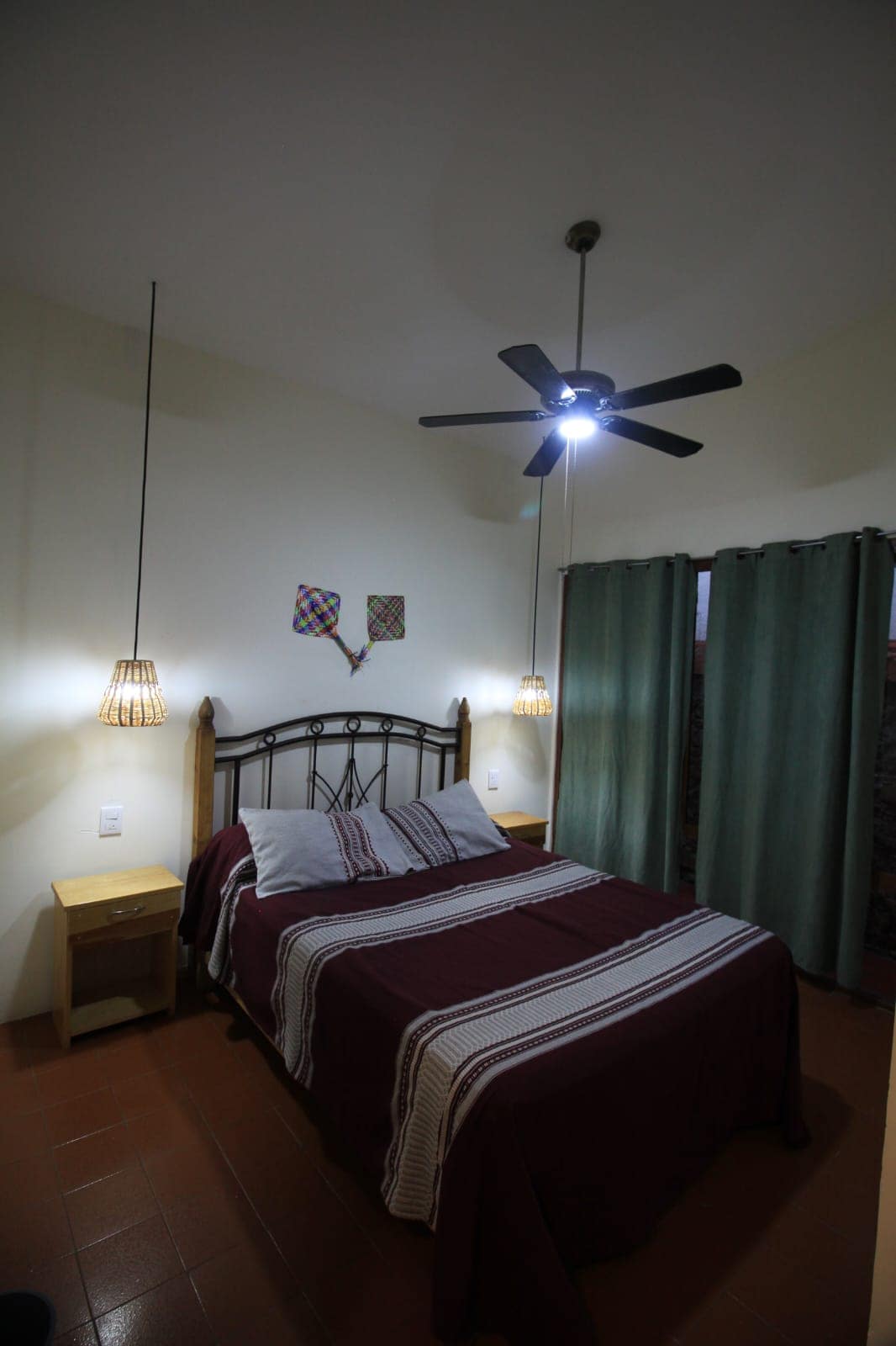 A cozy bedroom features a spacious bed with a maroon and white striped blanket, complemented by decorative pillows. Two woven hanging light fixtures illuminate the space, and a ceiling fan provides airflow. Green curtains frame the window, while two small nightstands are positioned beside the bed.