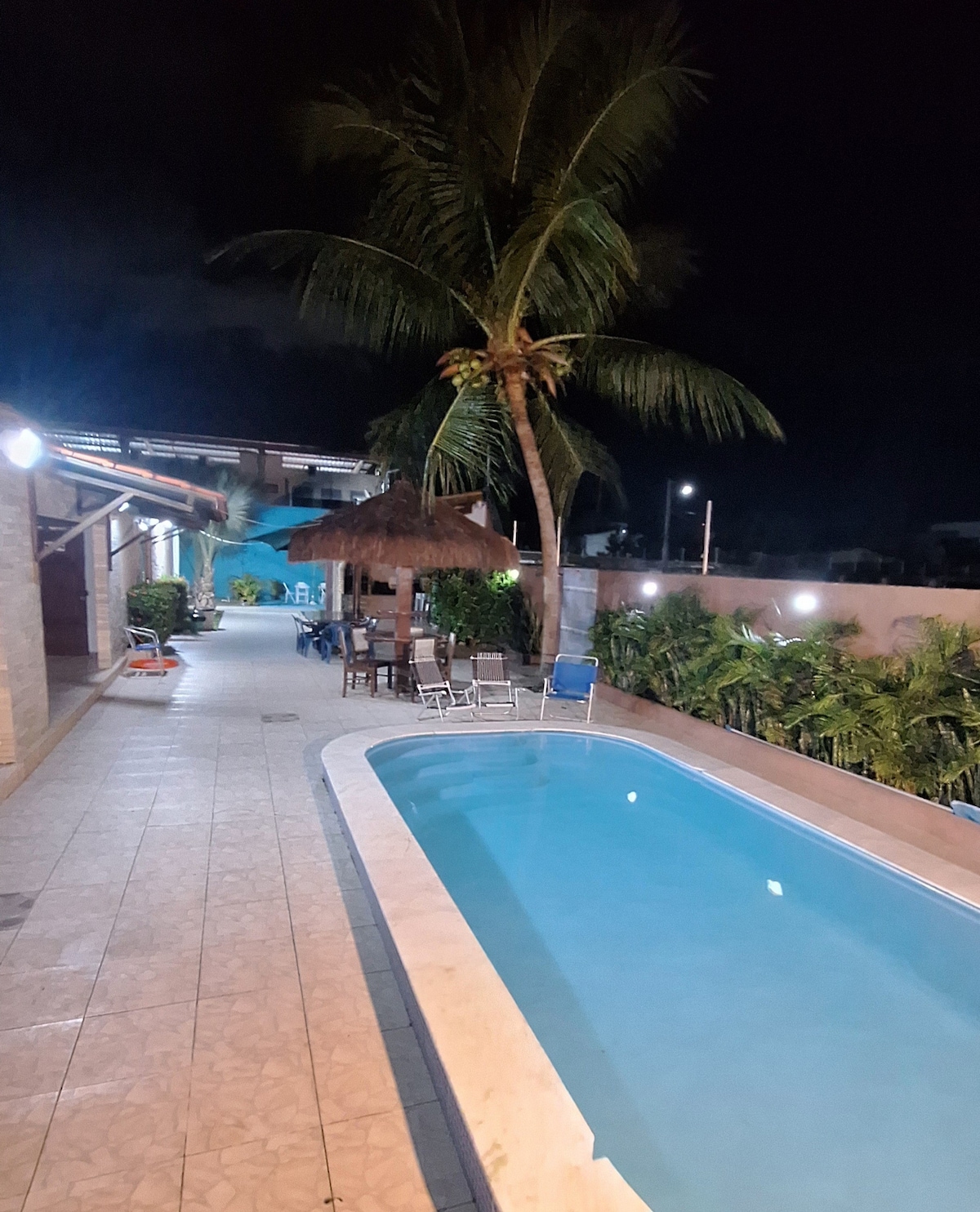 A well-lit pool is seen beside a palm tree, surrounded by a tiled pathway. Lounge chairs and a thatched gazebo are positioned nearby, with greenery lining the area. The evening sky creates a calm atmosphere, highlighting the inviting space for relaxation.