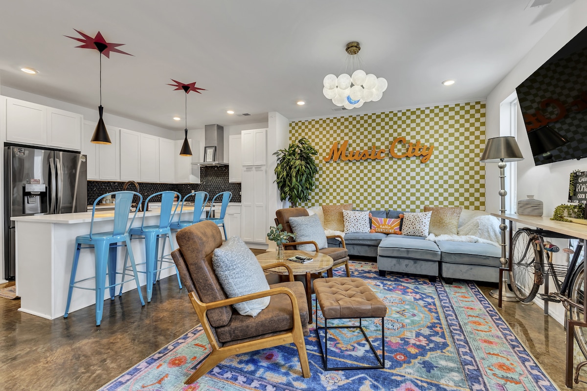 An open living area showcases a comfortable seating arrangement with a mix of chairs and a sofa set against a backdrop of vibrant decor. A feature wall with 'Music City' lettering is highlighted, while modern bar stools and a well-equipped kitchen are visible in the background.