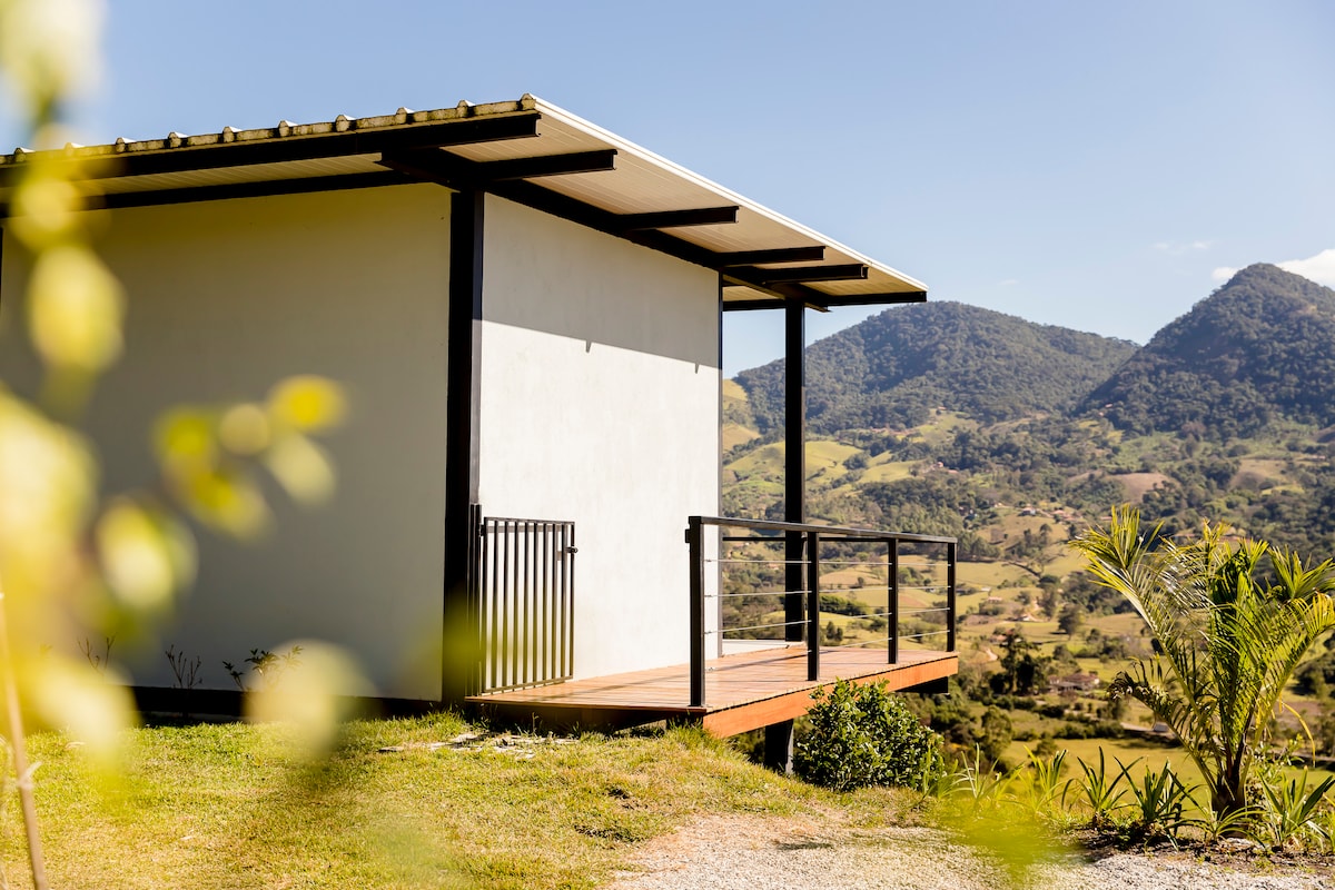 The exterior of a private chalet is shown, featuring a wooden deck that offers views of the surrounding mountains. The building is positioned on a slight elevation, with a clean, modern design and large windows allowing for natural light. Greenery is visible in the foreground.